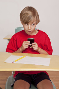 A young boy sitting at his desk in class trying to sneak a text on his cell phone as he peeks out of the corner of his eyes looking for the teacher.